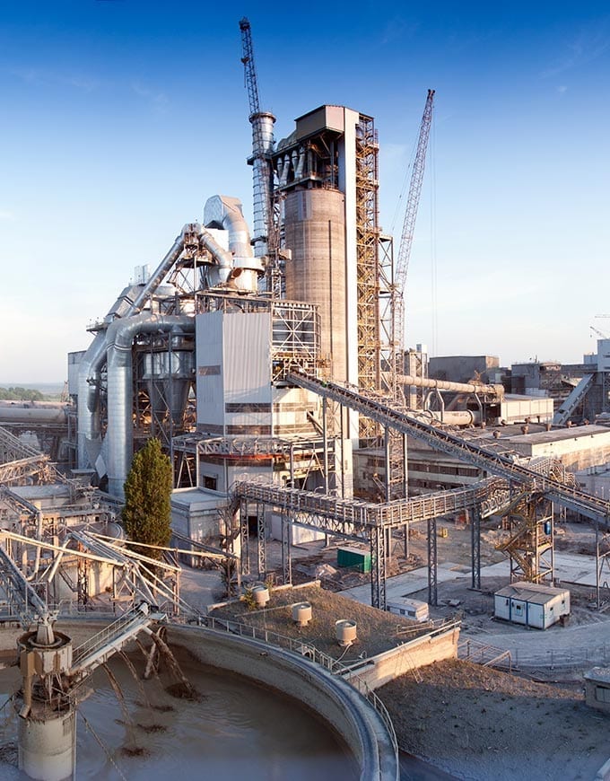 Large industrial cement plant with tall silos, conveyor belts, metal structures, and cranes under a blue sky.