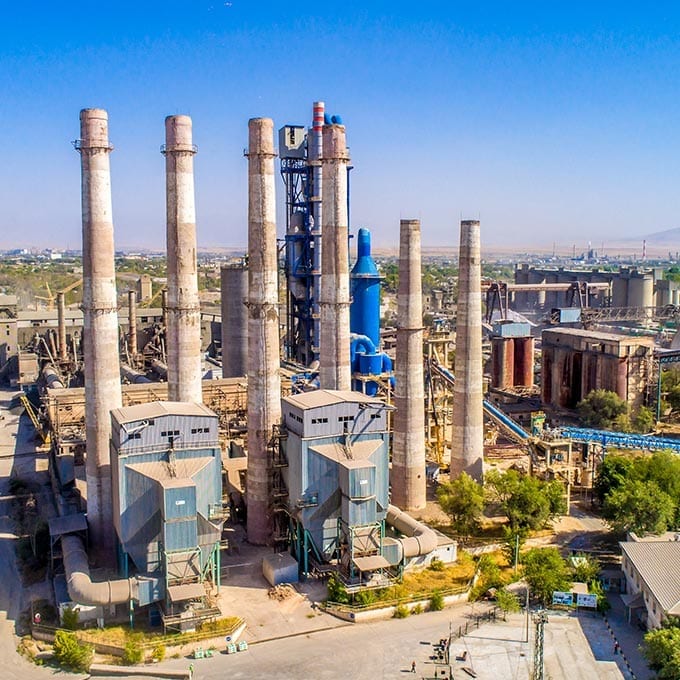 Aerial view of a large industrial cement plant with multiple tall concrete chimneys, industrial buildings, and machinery under a clear blue sky.