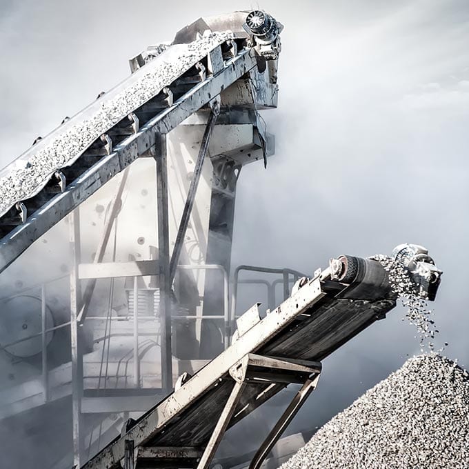 Two conveyor belts transport and deposit gravel at an industrial site, with dust and debris visible in the air.