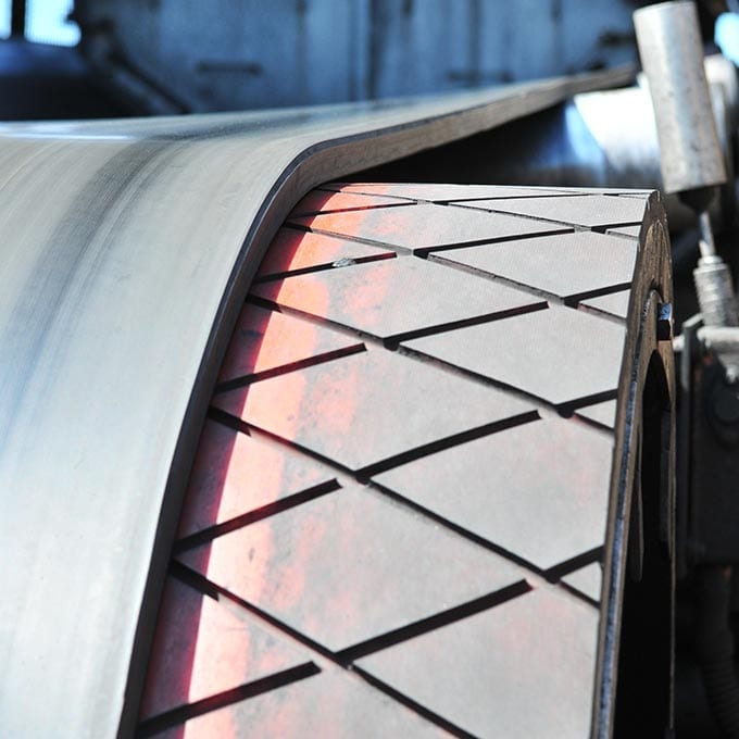Close-up of an industrial conveyor belt running over a patterned drive pulley, with a red stripe visible along the edge of the pulley surface.