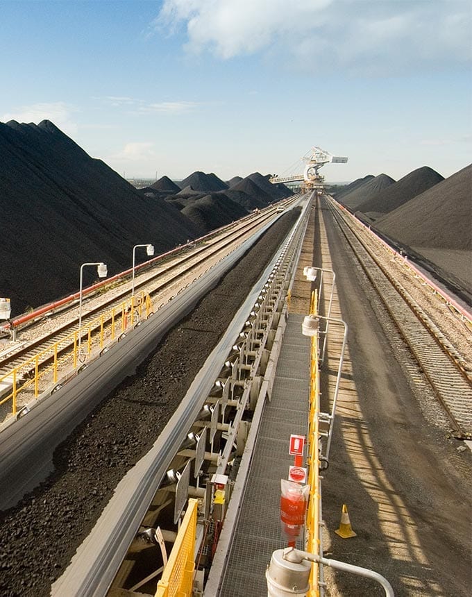 Conveyor belts transport coal alongside large stockpiles at an industrial mining facility under a partly cloudy sky.