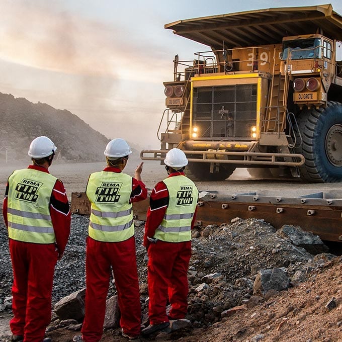 Three workers in safety gear stand on a rocky site, signaling to a large yellow mining dump truck approaching them.