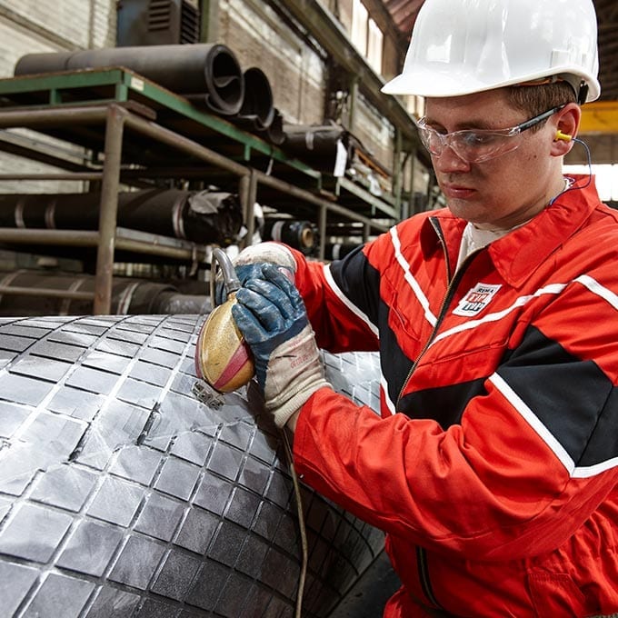 A worker in safety gear grinds a large, checkered metal cylinder in an industrial setting with shelves and pipes in the background.