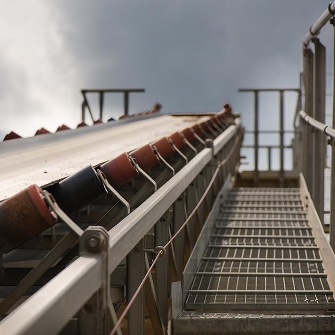 A metal conveyor belt is positioned next to a grated metal walkway, both leading upward toward an overcast sky.
