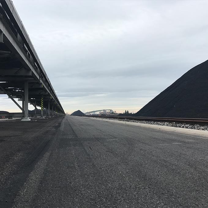 Empty industrial roadway with a conveyor structure on the left and large piles of black material, likely coal, on the right under an overcast sky.