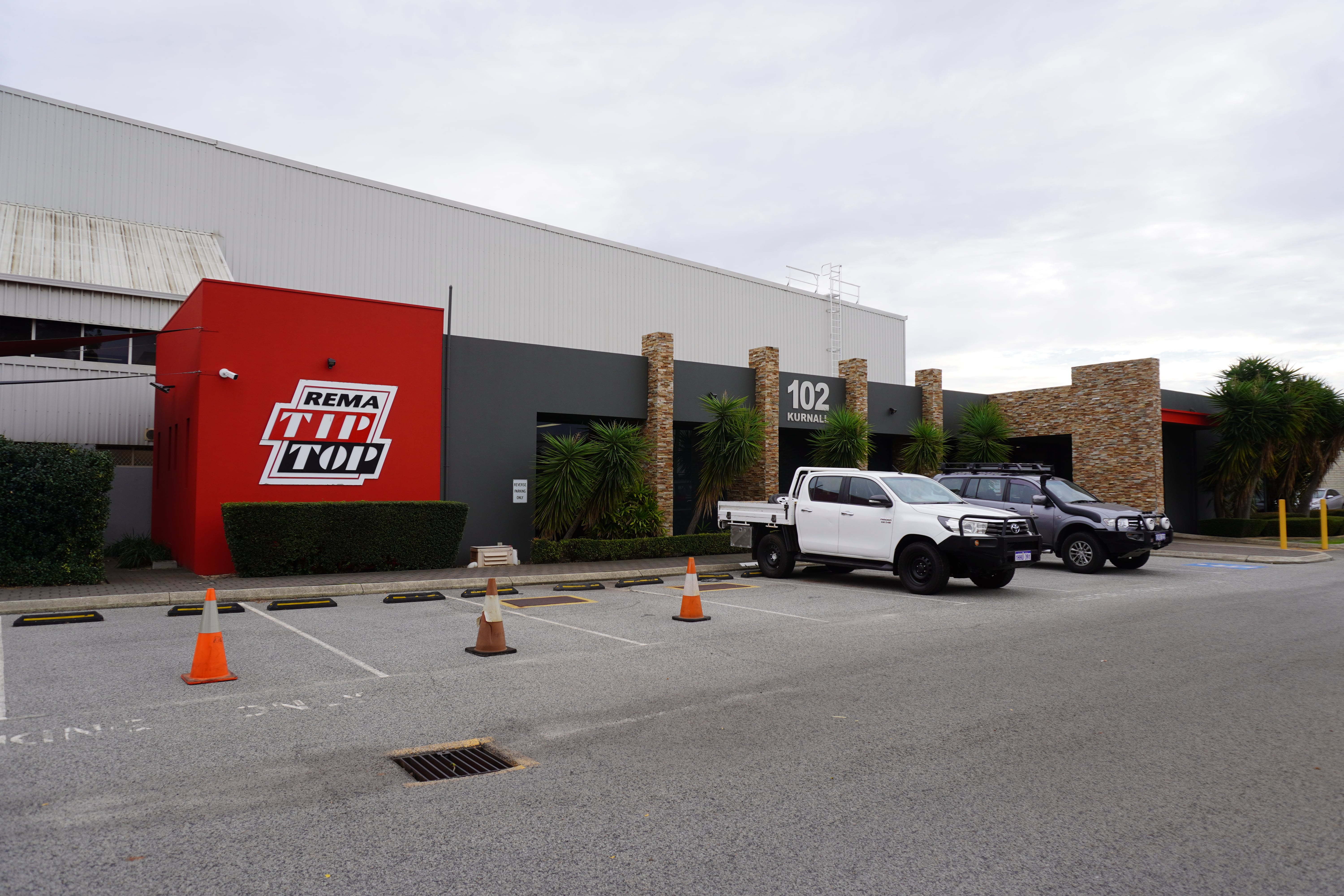 Two parked utility vehicles in front of a REMA Tip Top building with a red facade, labeled 102, and orange traffic cones in the parking lot.