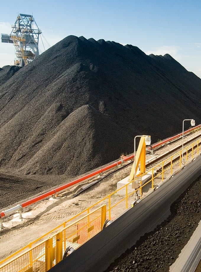 Large mounds of coal piled in an industrial facility, with conveyor belts and machinery in the foreground under a clear sky.