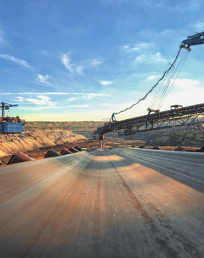 A wide conveyor belt transports materials at an open-pit mining site under a blue sky, with large machinery and terraces visible in the background.
