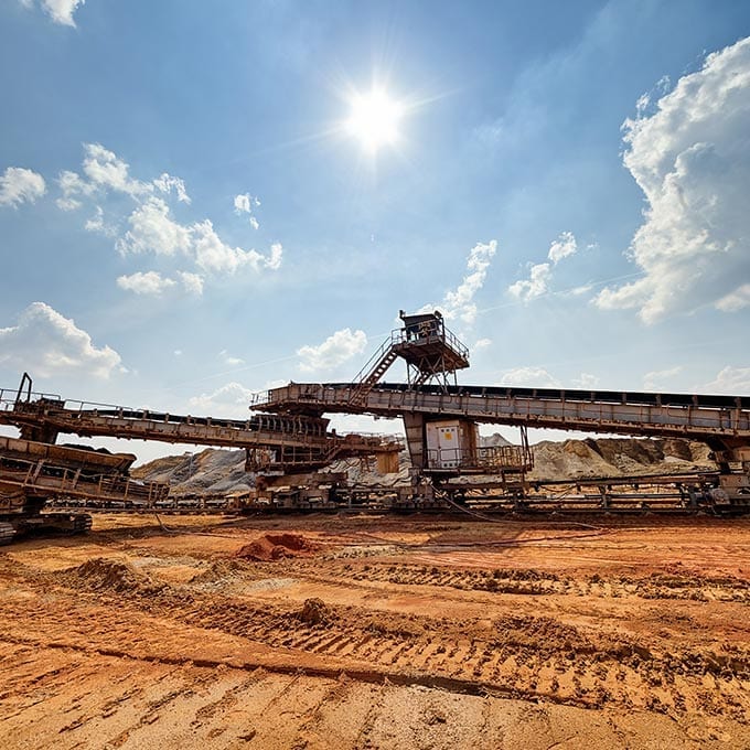 Large mining equipment operates on a sandy site under a bright sun and blue sky, with conveyor belts and machinery visible in the scene.
