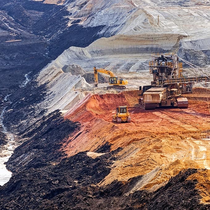 Large excavators and machinery operate on terraced layers of an open-pit mine, revealing exposed earth in varying colors.