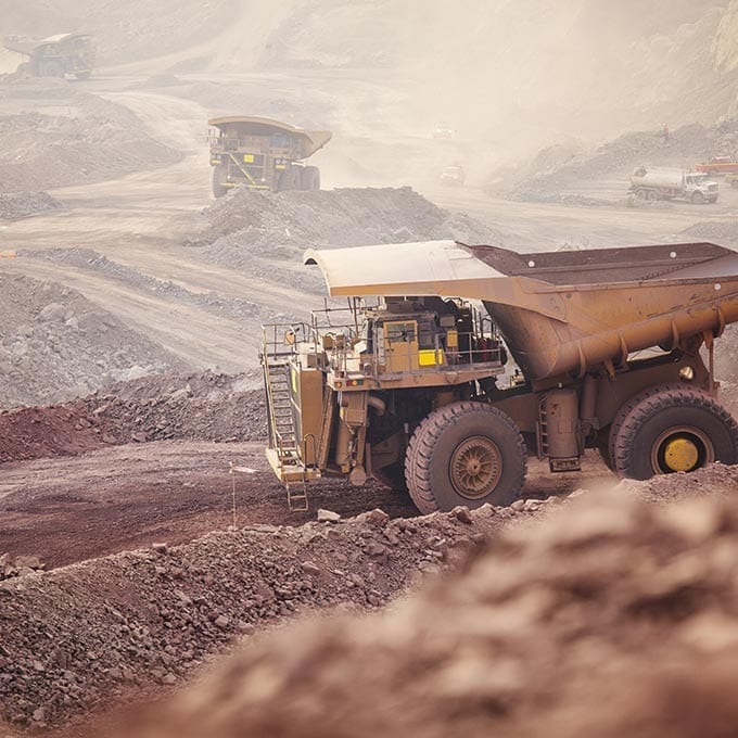 Large mining dump trucks operate on a dirt road in an open-pit mine, surrounded by piles of excavated earth and rocks.