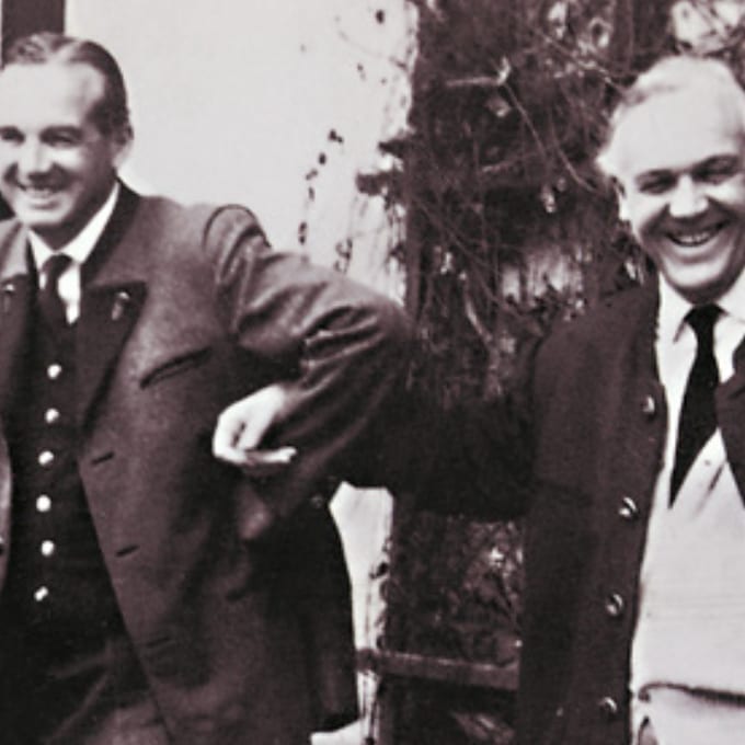 Two men in suits and coats stand outdoors, smiling and holding arms. The image is in black and white.
