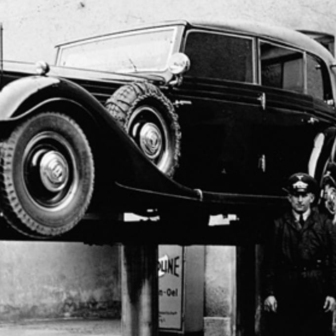 A vintage car is elevated on a hydraulic lift as a uniformed attendant stands beside it in a garage setting.