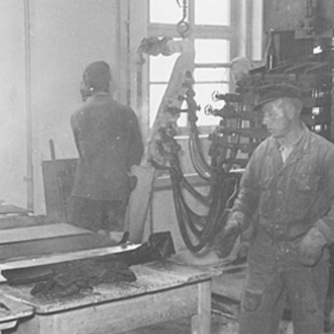 Two men work in an industrial factory setting with machinery, tables, and tools visible; one faces away, while the other is in the foreground wearing work clothes and gloves.