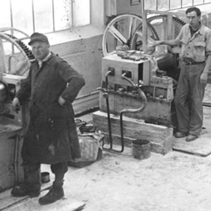 Two men stand in an industrial workshop near large mechanical equipment and wheels, with tools and machinery visible in the background.
