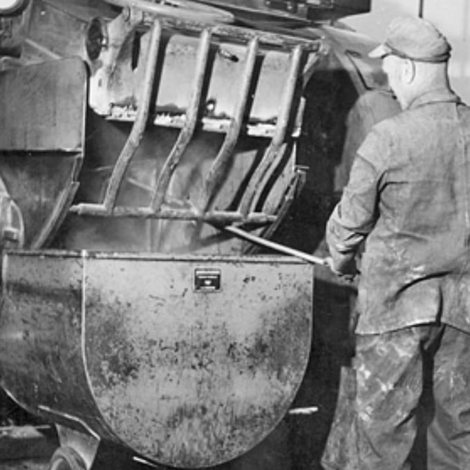 A worker wearing protective clothing stands beside a large industrial mixing machine, using a tool to stir or manage the contents inside the metal container.
