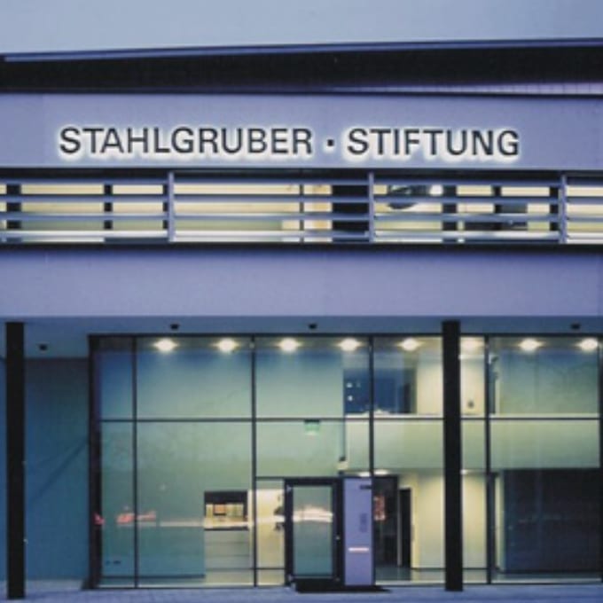 Modern building facade with large glass windows and STAHLGRUBER · STIFTUNG sign above the entrance, illuminated at night.