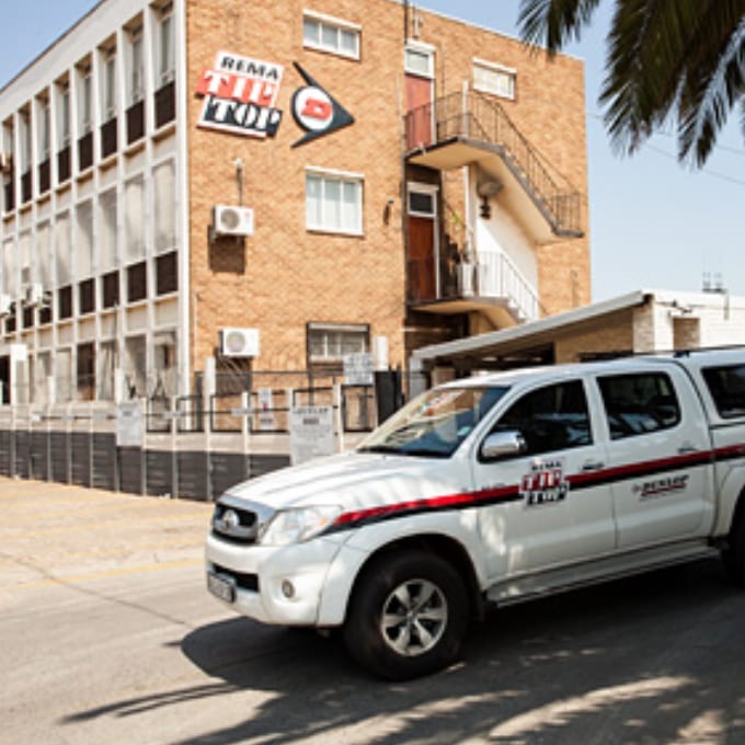 A white pickup truck with REMA TIP TOP branding parked in front of a brick office building with the same logo on the exterior wall.