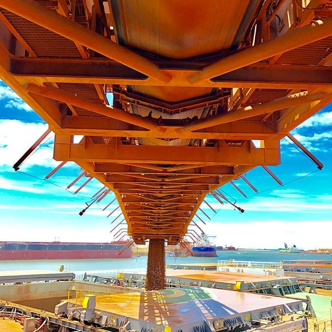 View from below of a large industrial conveyor system loading bulk material onto a docked ship, with blue sky and distant ships visible in the background.