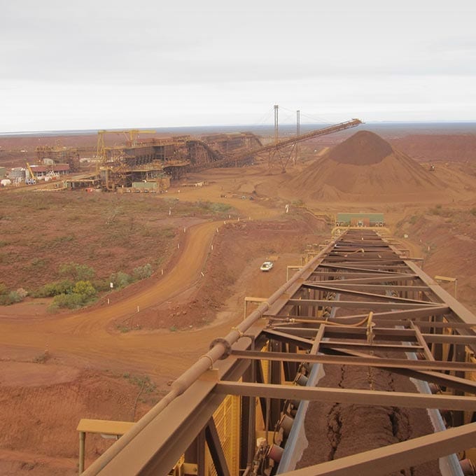 An iron ore mining facility with conveyor belts, large piles of extracted ore, and industrial equipment set in a reddish, barren landscape under a cloudy sky.