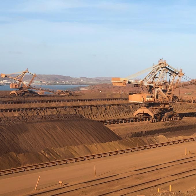 Large industrial mining machines operate on a vast, dusty site with mounds of earth and conveyor belts; hills and water are visible in the background under a blue sky.