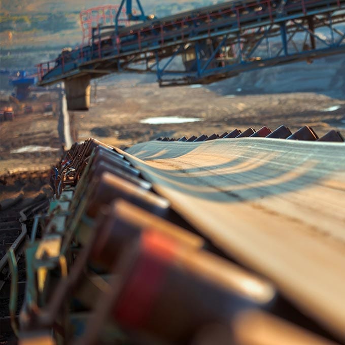 A close-up view of an industrial conveyor belt system transporting material at a mining or construction site, with other equipment visible in the background.