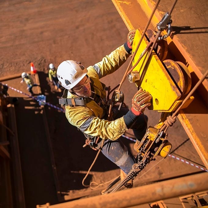 Worker in safety gear climbs industrial structure using ropes and harness, with colleagues and caution tape visible on the ground below.