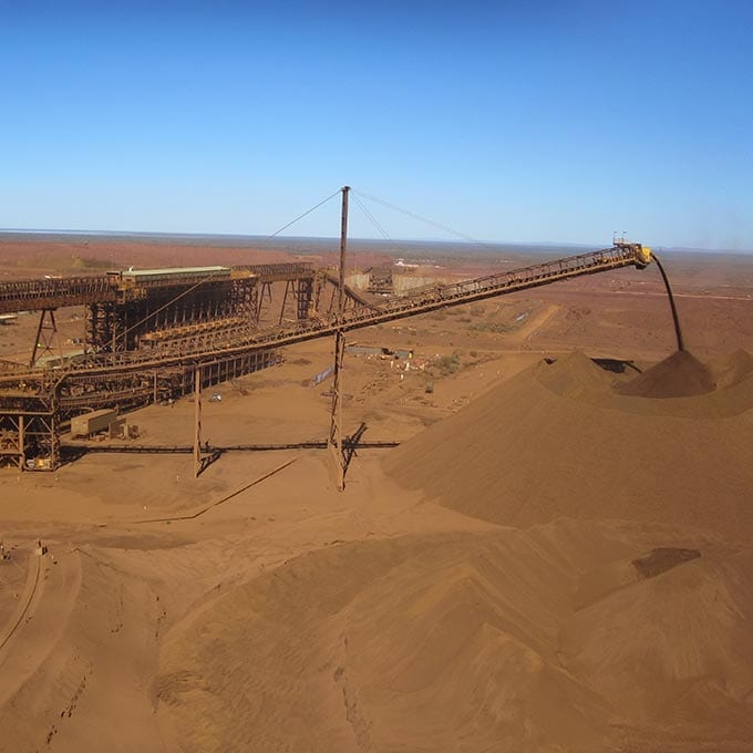 A large conveyor system deposits iron ore onto a massive pile at an open-pit mining site under a clear blue sky.