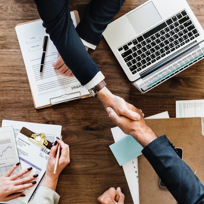 Three people at a desk with documents and a laptop; two people are shaking hands while a third takes notes on a clipboard.