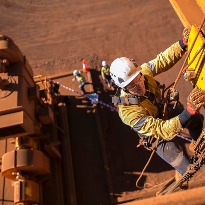 A worker wearing protective gear climbs industrial equipment at a mining site, while three other workers stand in the background on the ground.