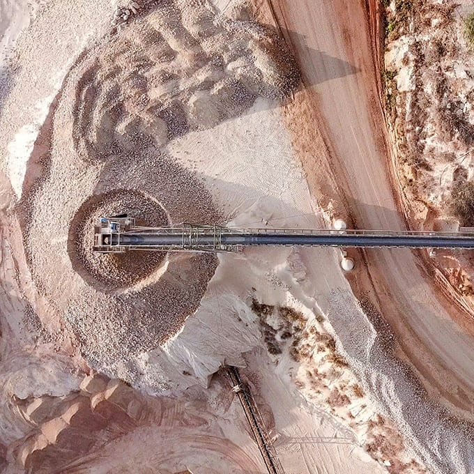 Aerial view of an industrial conveyor belt depositing gravel onto a large circular pile at a mining or quarry site.