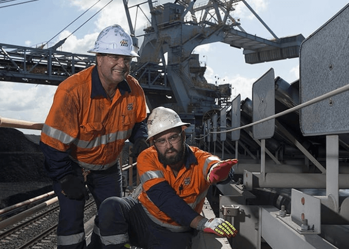 Two workers in orange safety gear and helmets inspect machinery on an industrial site.