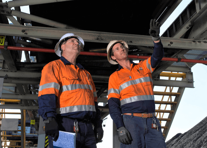 Two workers in orange safety gear and hard hats inspect industrial machinery. One points upward while the other holds a clipboard.