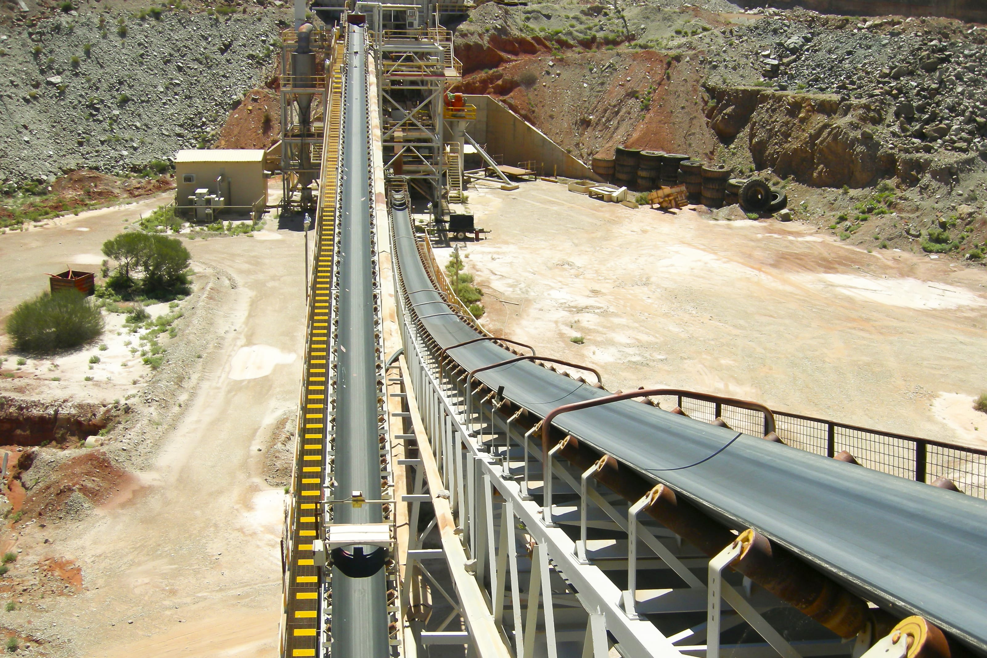 An elevated conveyor belt system at an open-pit mine, transporting materials toward processing facilities surrounded by rocky terrain and arid landscape.