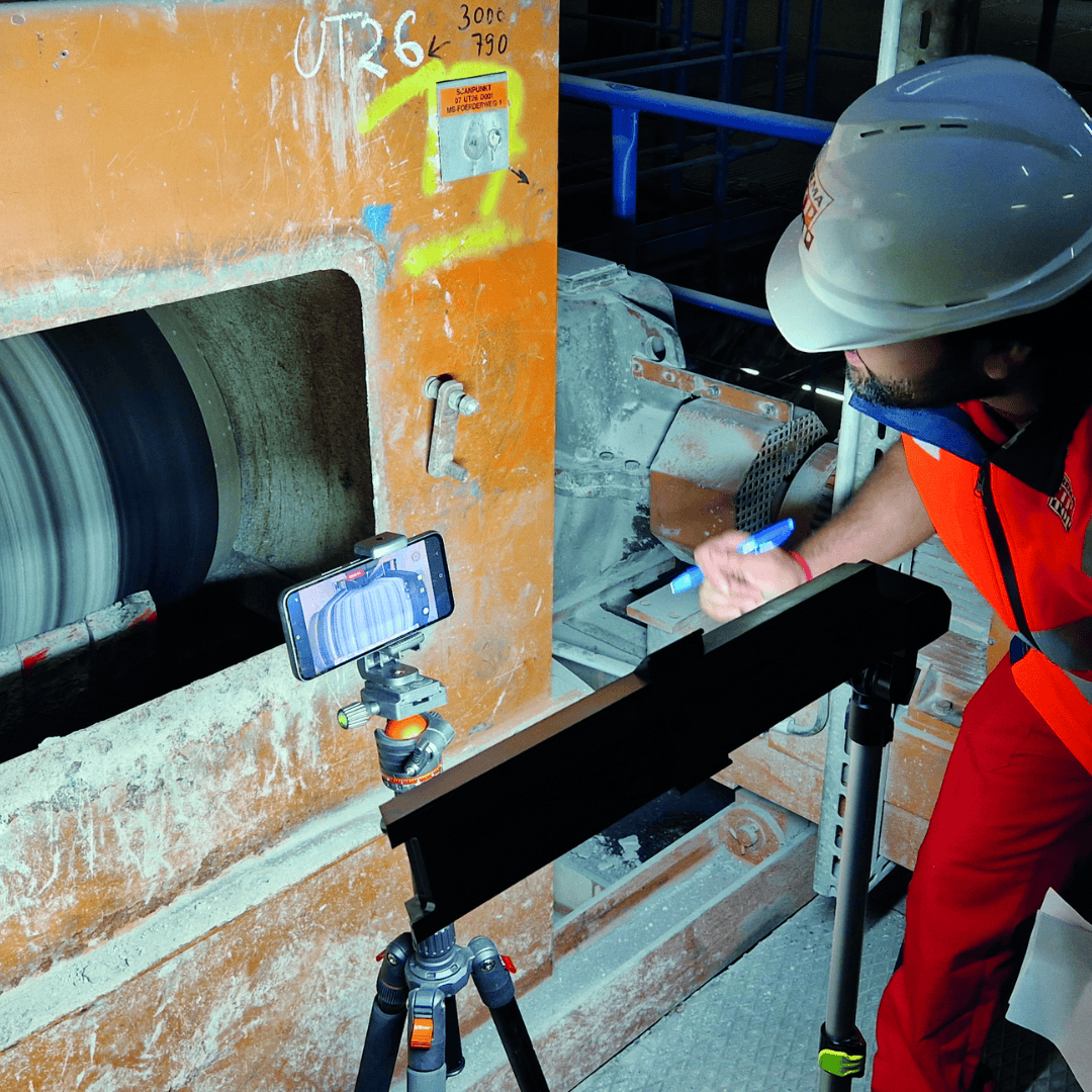 A person in safety gear records data on a notepad while a phone on a tripod films a rotating industrial machine inside a factory.