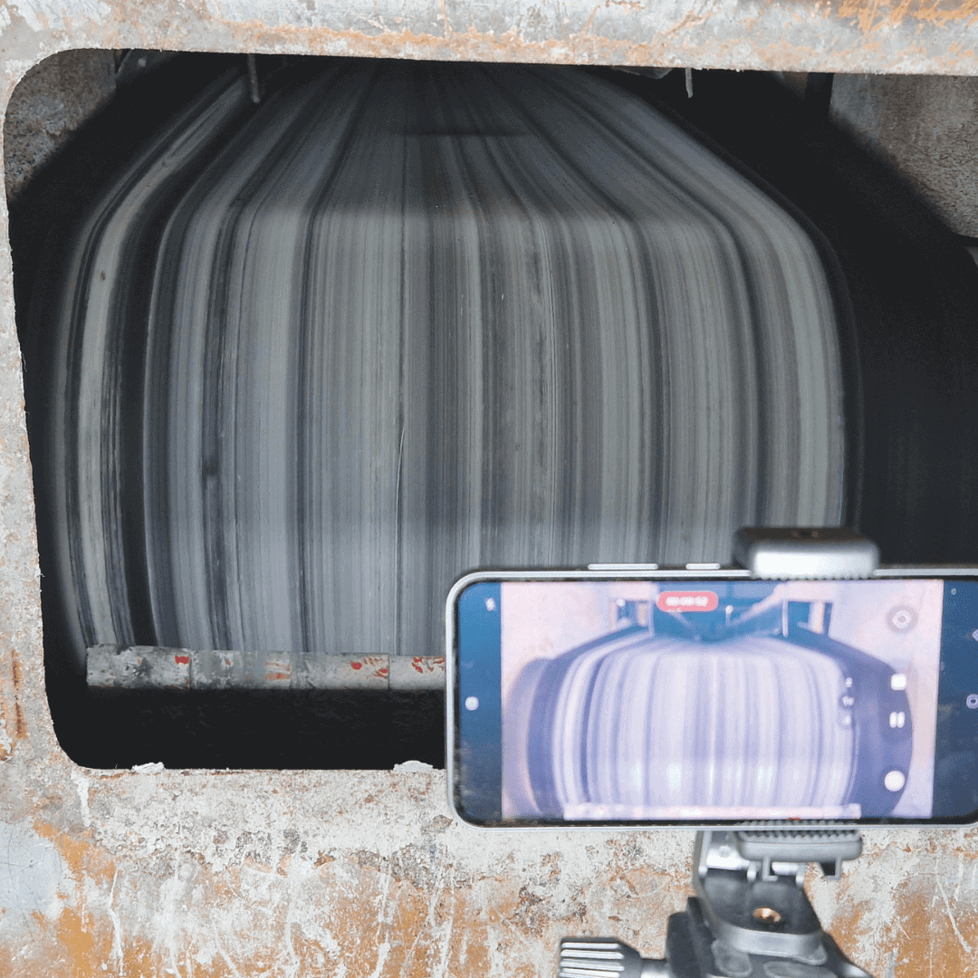 A smartphone on a tripod records a large industrial machine with a spinning striped cylindrical component through a rectangular opening.