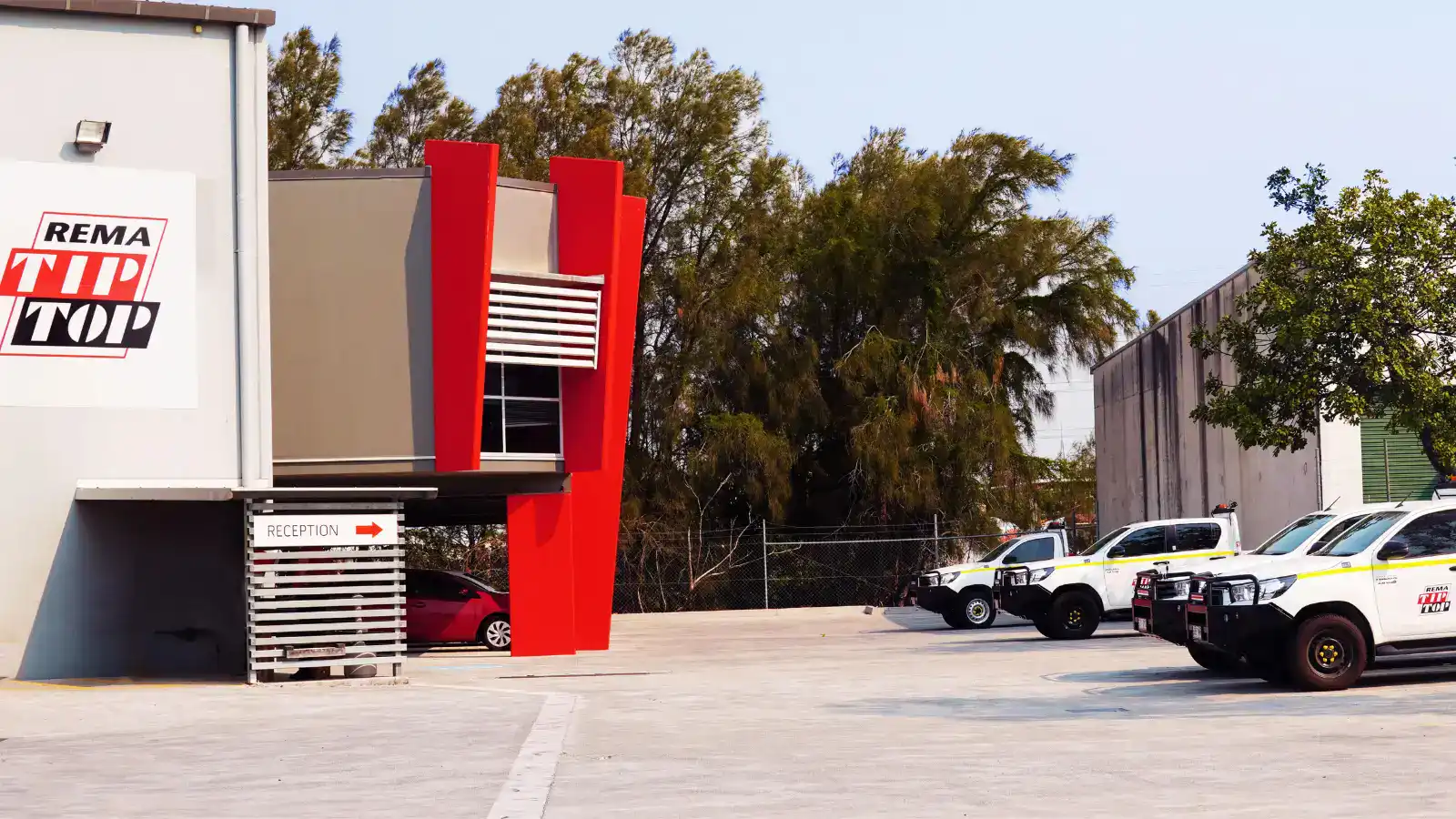A commercial building with a “REMA TIP TOP” sign, a visible reception area, and several parked utility vehicles in an outdoor lot.