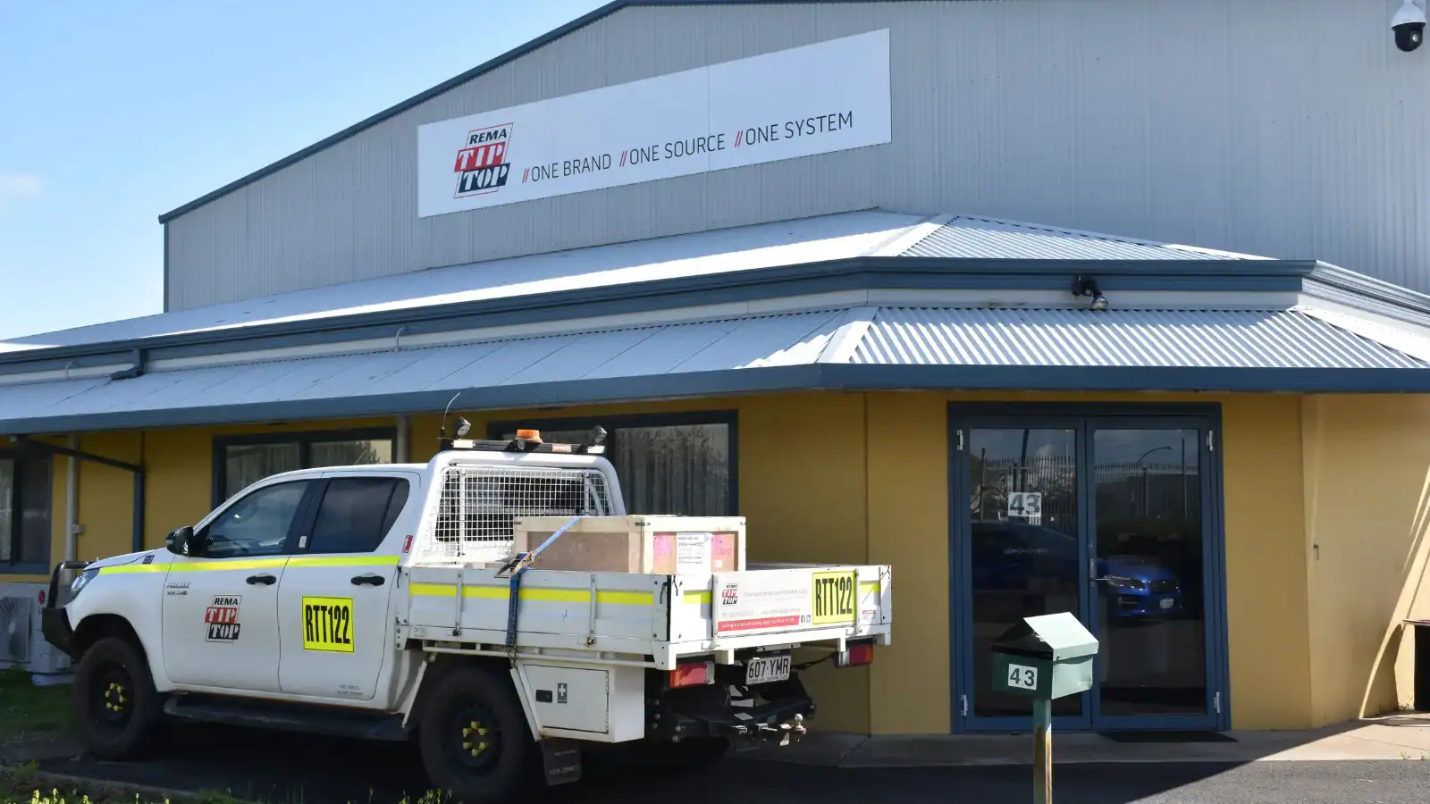 A white utility vehicle is parked outside a commercial building with a sign reading “TTi / One Brand / One Source / One System.” The building has a corrugated metal roof and yellow walls.
