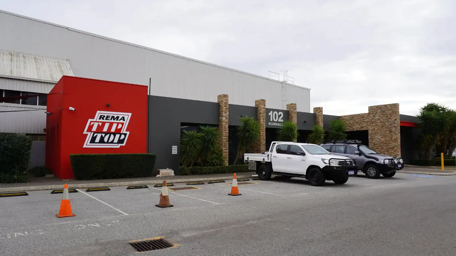 Two parked utility vehicles in front of a REMA Tip Top building with a red facade, labeled 102, and orange traffic cones in the parking lot.